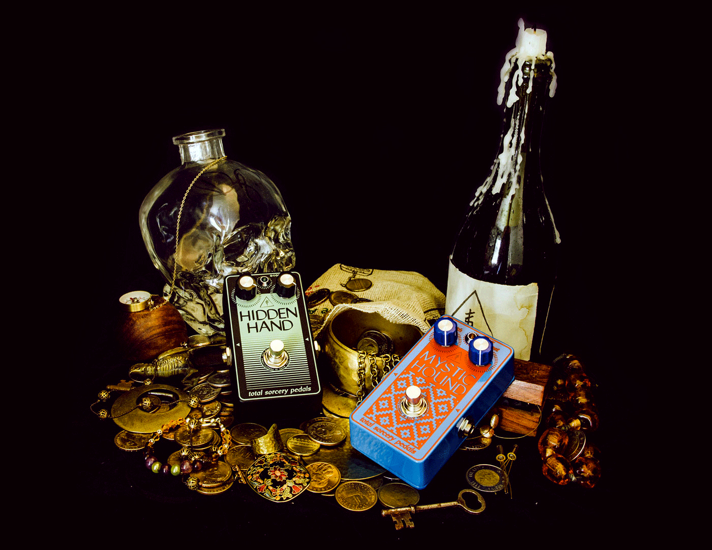 Two guitar pedals lay on a treasure trove of coins. A wine bottle and a crystal skull